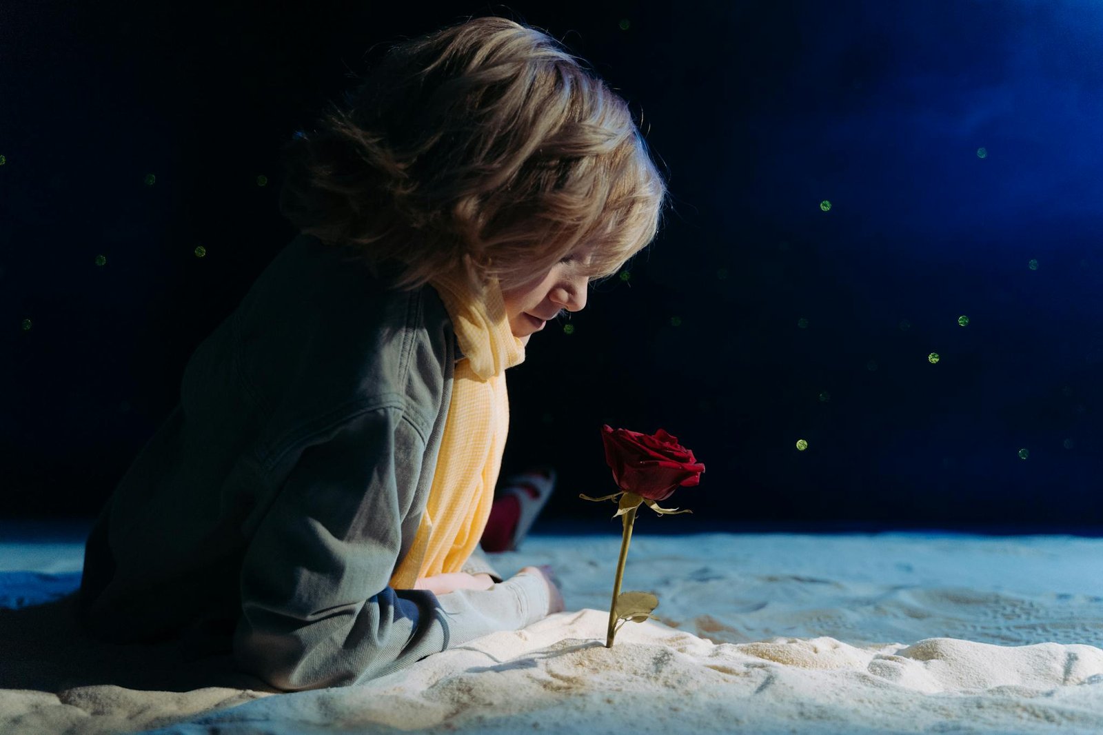 A child in cosplay as the Little Prince gazing at a single red rose against a starry night backdrop.