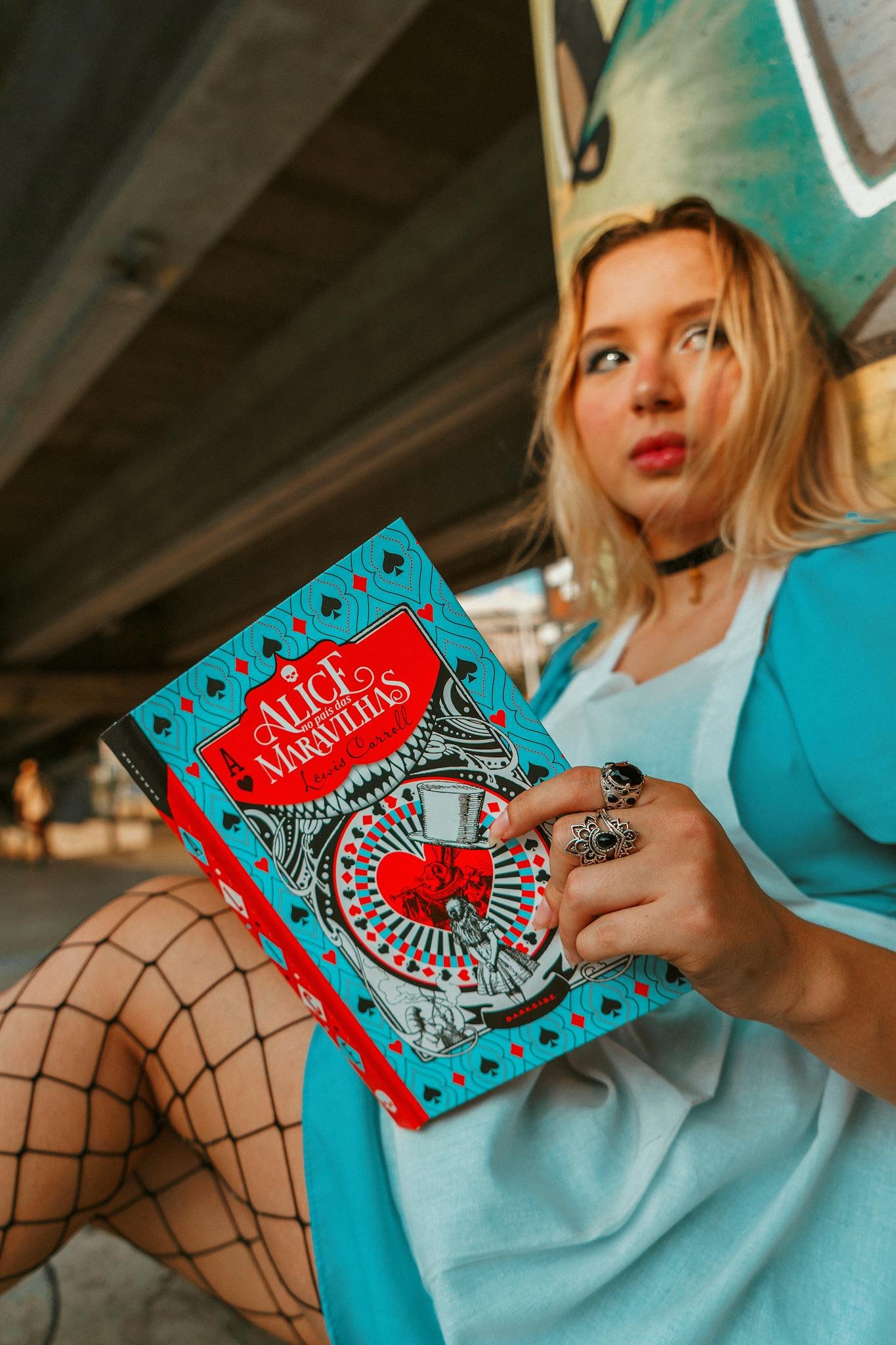 Woman dressed as Alice reading under an urban bridge.